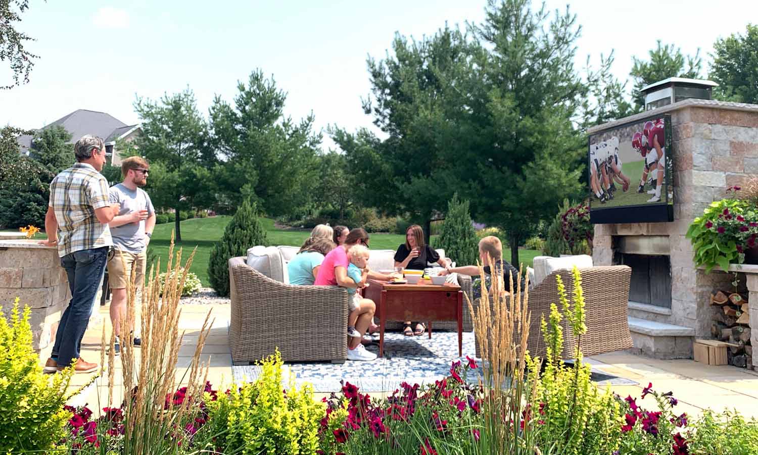 Group of friends sit outdoors while watching football on an outdoor TV mounted above a fireplace