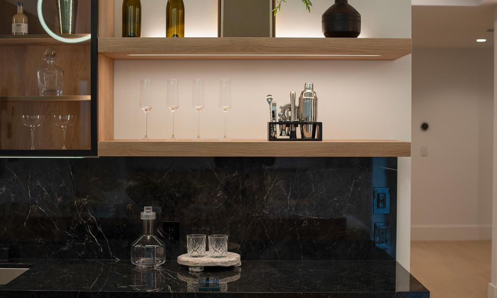 Modern bar area with backlit wooden shelves holding glassware and bar tools above a dark marble counter
