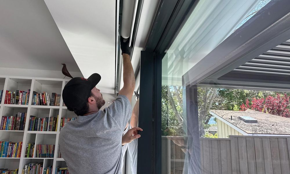 Technician adjusting a ceiling-recessed roller shade inside a modern home with glass wall and bookshelves