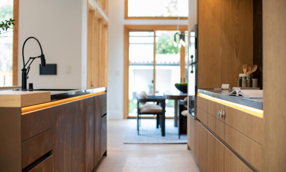 View down a modern kitchen galley with integrated LED lighting under countertops and a dining table in the distance