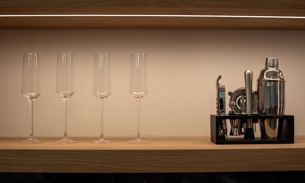 Close-up of champagne flutes and a bar tool set on a wooden shelf with subtle LED lighting