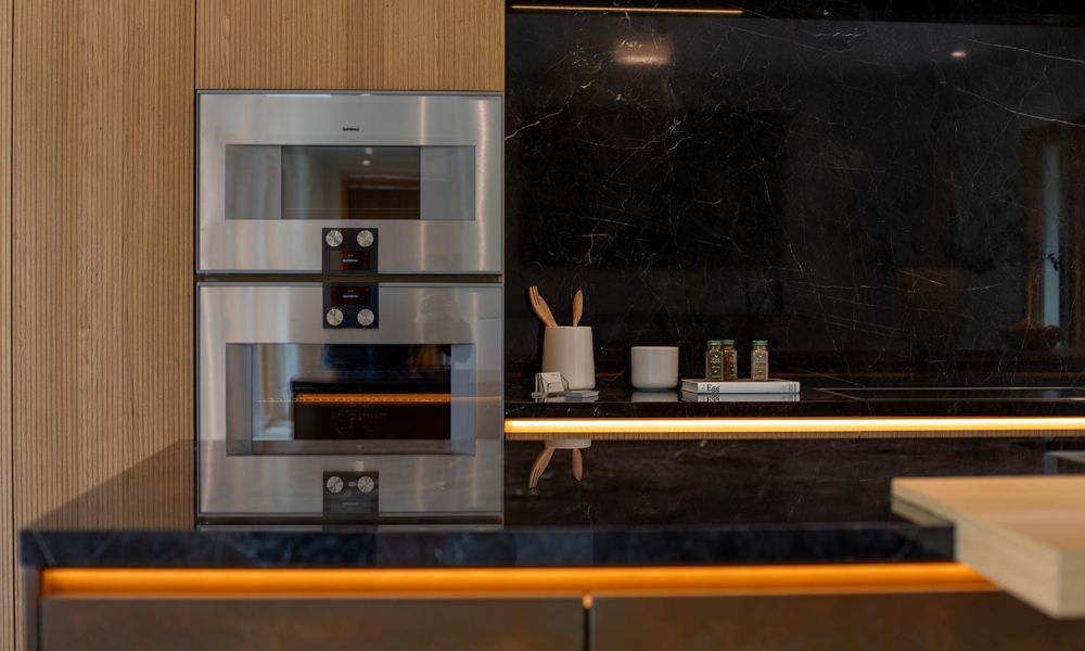 Close-up of stacked stainless steel ovens beside a dark marble backsplash and LED strip lighting