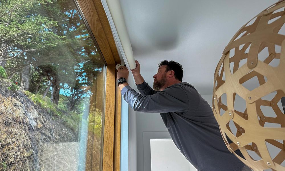 Technician standing on a ladder installing a roller shade over a tall window framed with wood