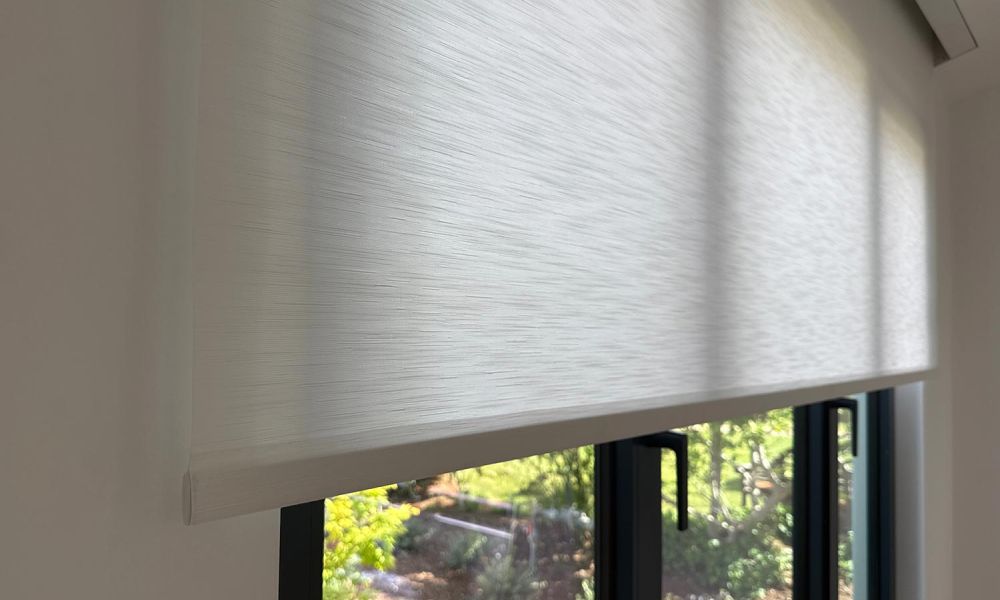 White roller shade partially lowered over black-framed windows with a garden view outside